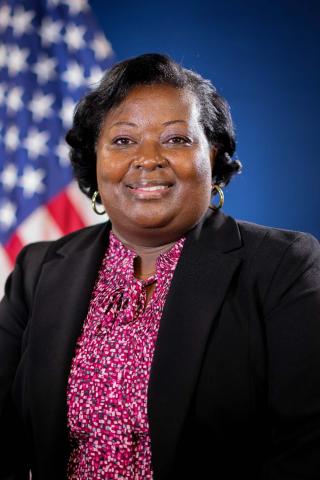 Woman in a black blazer and pink blouse in front of the American flag and a blue background