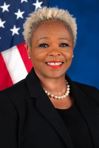Portrait of a smiling woman with short, blonde hair. She is dressed in black and over her left shoulder is the American flag.