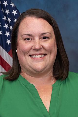 Female with shoulder length straight brown hair wearing a green top positioned in front of the American flag.
