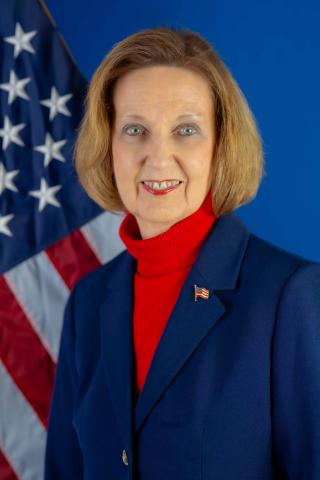 Ruth Mustard poses for a photo with a blue backdrop and a US flag over her right shoulder