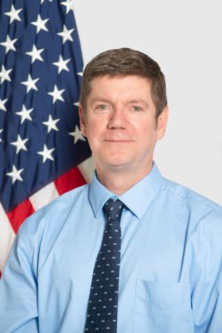 Acting associate director Casey Thayer in blue short and blue tie with US flag over right shoulder.