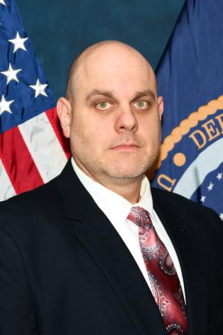 Man standing in a black suit in front of american flag and veteran affairs flag