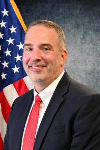 Dr. Thom Strom: a smiling, white man sitting in front of an American Flag wearing a black suit, white shirt and red tie.