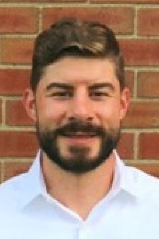 Man with dark hair and beard with light button up shirt.  Background is a brick wall.