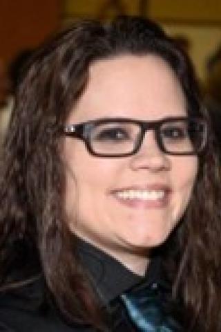 Woman with dark wavy hair, dark rimmed eyeglasses with dark colored shirt.