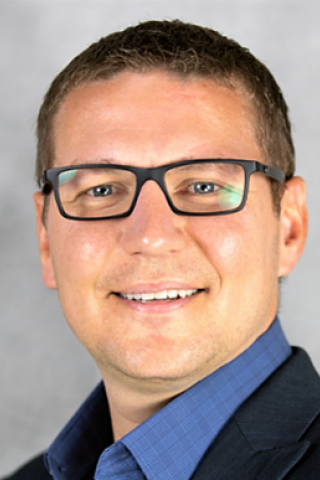 Man with dark-framed glasses, medium-navy blue button-down shirt and dark blue suit jacket.