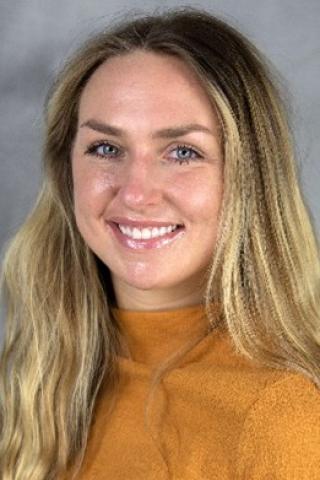 Woman with long blonde hair, wearing an orange blouse.  Background is grayish cloud-like backdrop.