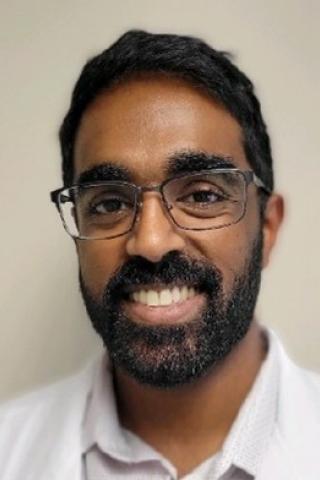 man with black hair, beard and mustache, wearing glasses and white button down polo-style shirt.  Background is a cream colored wall.