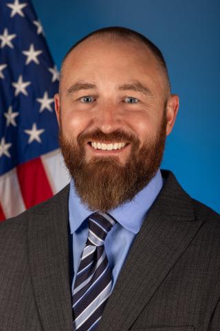 White man with brown beard wearing a blue shirt, grey suit coat, and blue and light blue striped tie, sitting in front of an American flag