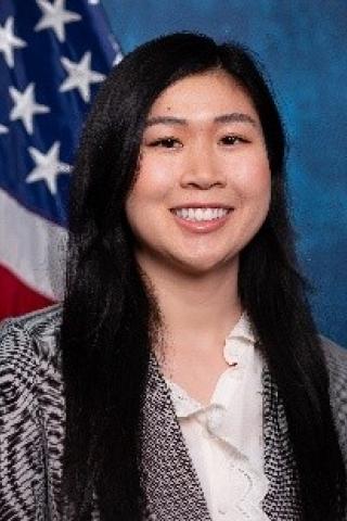 Young woman with long black hair, white blouse, black & white blazer.  Background is standard blue with American flag.