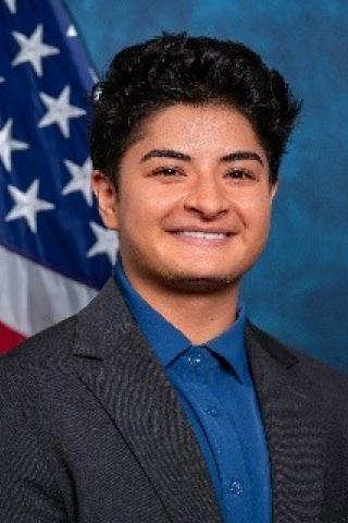 Young adult with black hair wearing blue shirt and a dark blue or black blazer.  Background standard blue with American flag.