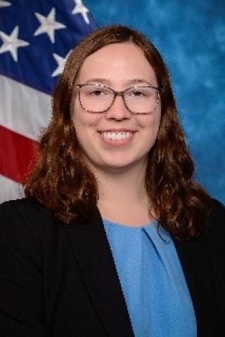 Young woman with dark hair, glasses, light blue blouse, dark blue or black blazer.  Background is standard blue with the American flag.
