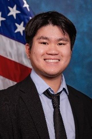 Young man with black hair, light blue shirt with dark blue/black tie and blazer.  Background is standard blue with American flag.