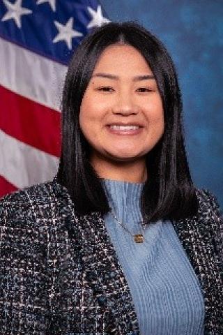 Young woman with black hair, light blue blouse, black and white tweed blazer, standard blue background with American Flag.