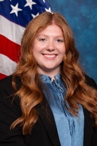 Young woman with long red hair, light blue blouse, dark blue / black blazer.  Background of standard blue with American flag.