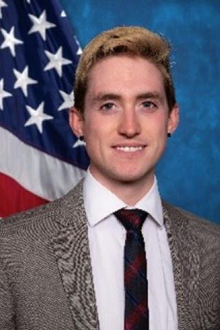 Young man with blond hair, white shirt, black tie and black and white tweed blazer.  Background standard blue with American flag