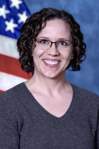 Woman, Caucasian, dark curly hair with eye glasses, wearing gray shirt,  Background is standard blue with American flag.