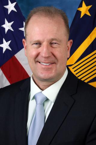 Caucasian male wearing white button down shirt, light blue tie, black suit coat.  Background is standard blue with American flag.