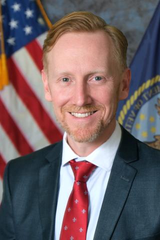 Man in blue suit and red tie with flags behind him.