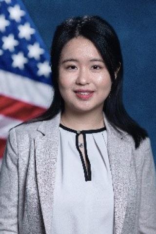 Young woman with black hair, white blouse, light gray blazer.  Background standard blue with American Flag.