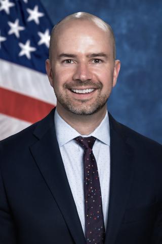 Caucasian, male, bald, white button-down shirt with dark colored tie and dark suit jacket.  Background is standard blue with American flag.