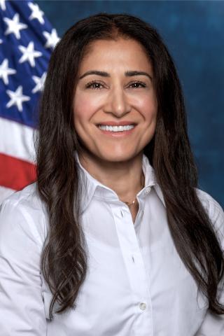 Woman of color, long dark hair, white button down shirt.  Background is standard blue with American flag.