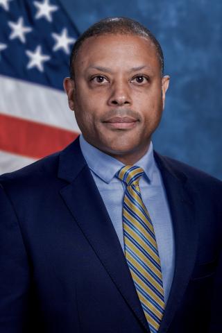 Male, person of color, light blue button down shirt, blue & yellow striped tie, navy blue suit coat.  Background is standard blue with American flag.