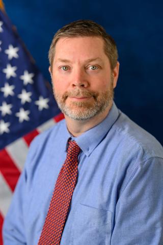 Chris Zell poses with a blue backdrop and U.S. flag in the background. 