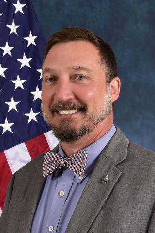 Dotts in grey blazer with blue shirt and patterned bowtie smiling in front of an American flag with a blue background