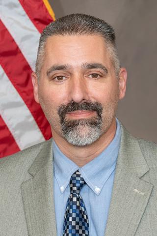 A man with a beard in a suit and tie stands in front of the US flag.
