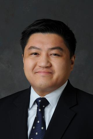 A man with short black hair wearing a suit and tie smiles at the camera.
