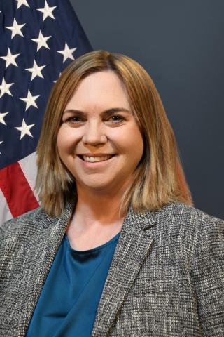 Woman with blonde hair wearing a blue shirt and gray blazer in front of an American flag.
