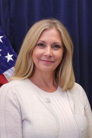 Woman with blonde hair smiling in front of a USA flag.