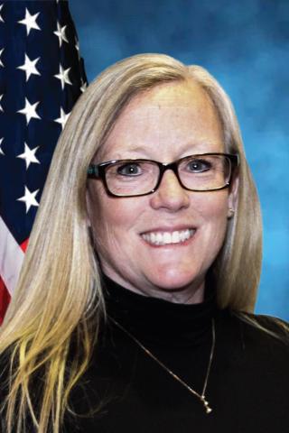 Portrait of a smiling woman with blonde hair wearing glasses and a black top, standing in front of a US flag.