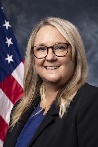 Blonde woman in black blazer and blue shirt with glasses, smiling in front of American flag.