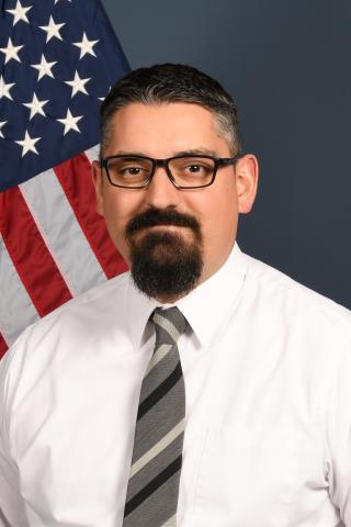 Man with glasses and beard wearing white shirt and gray tie in front of US flag.
