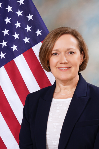 Woman in navy blazer and white shirt stands before American flag, smiling.
