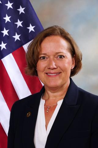 women with brown hair wearing a white shirt and black blazer in front of U.S. flag