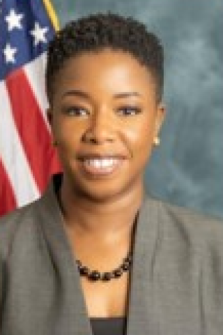 A woman with short hair and a gray blazer smiles in front of an American flag.