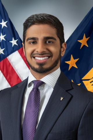 Man in suit and purple tie standing in front of US and DHS flags.