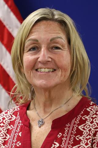 Portrait of smiling woman with blonde hair, red shirt, and silver necklace. American flag in background.