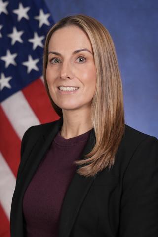 Woman with blonde hair in a black blazer and maroon top in front of a US flag.