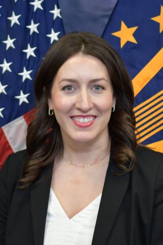 A smiling woman in a black blazer and white shirt stands in front of flags.