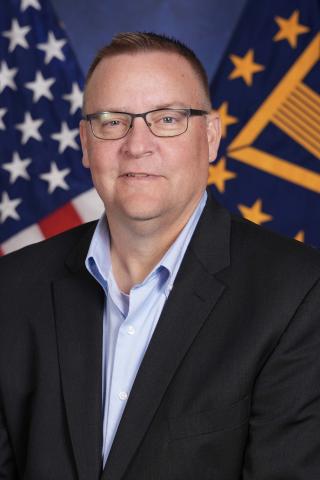 Man in black suit and blue shirt standing in front of American and Indiana flags.