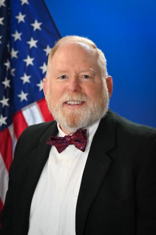 A smiling older man in a black suit with a bowtie stands in front of an American flag.