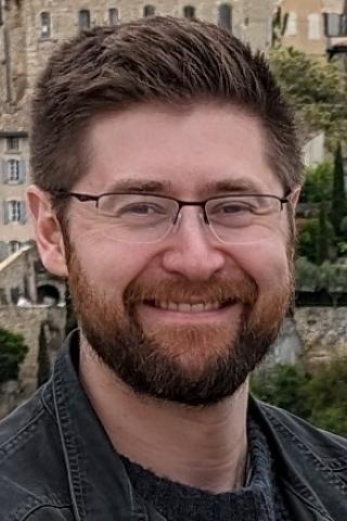 Photo of man with brown hair, brown beard, and glasses smiling in front of a town.