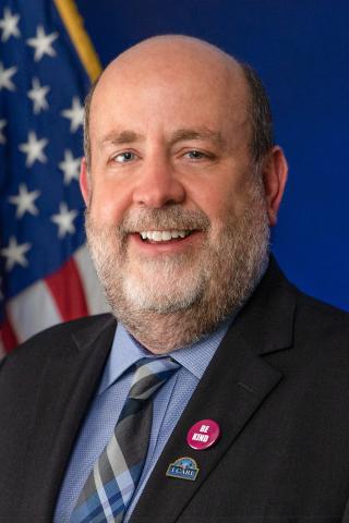 Portrait of a bald man with a beard, wearing a suit and tie, smiling against a blue background with an American flag.
