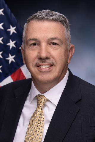Acting Associate Director Grady posing in front of the American Flag in a black jacket, white dress shirt, and yellow tie with blue circles