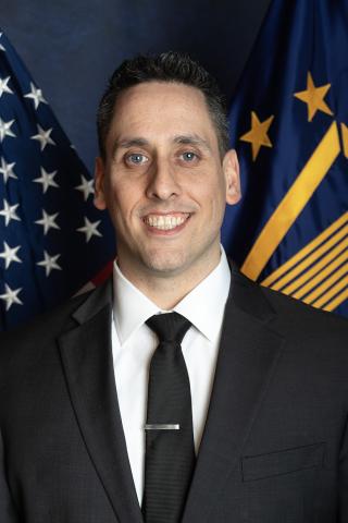 Man in black suit and tie smiling in front of American and Air Force flags.