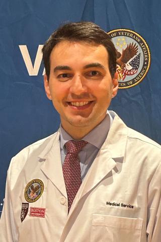 A smiling man in a white lab coat with a tie stands in front of a blue backdrop.